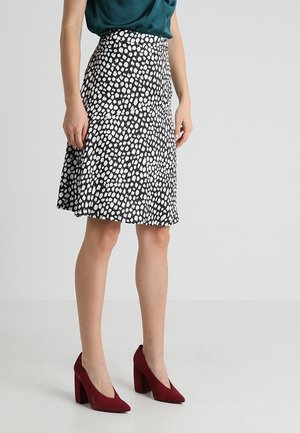 Femme portant une jupe mi-longue noire à pois blancs, un haut vert foncé et des chaussures à talons rouges, debout sur un sol clair.