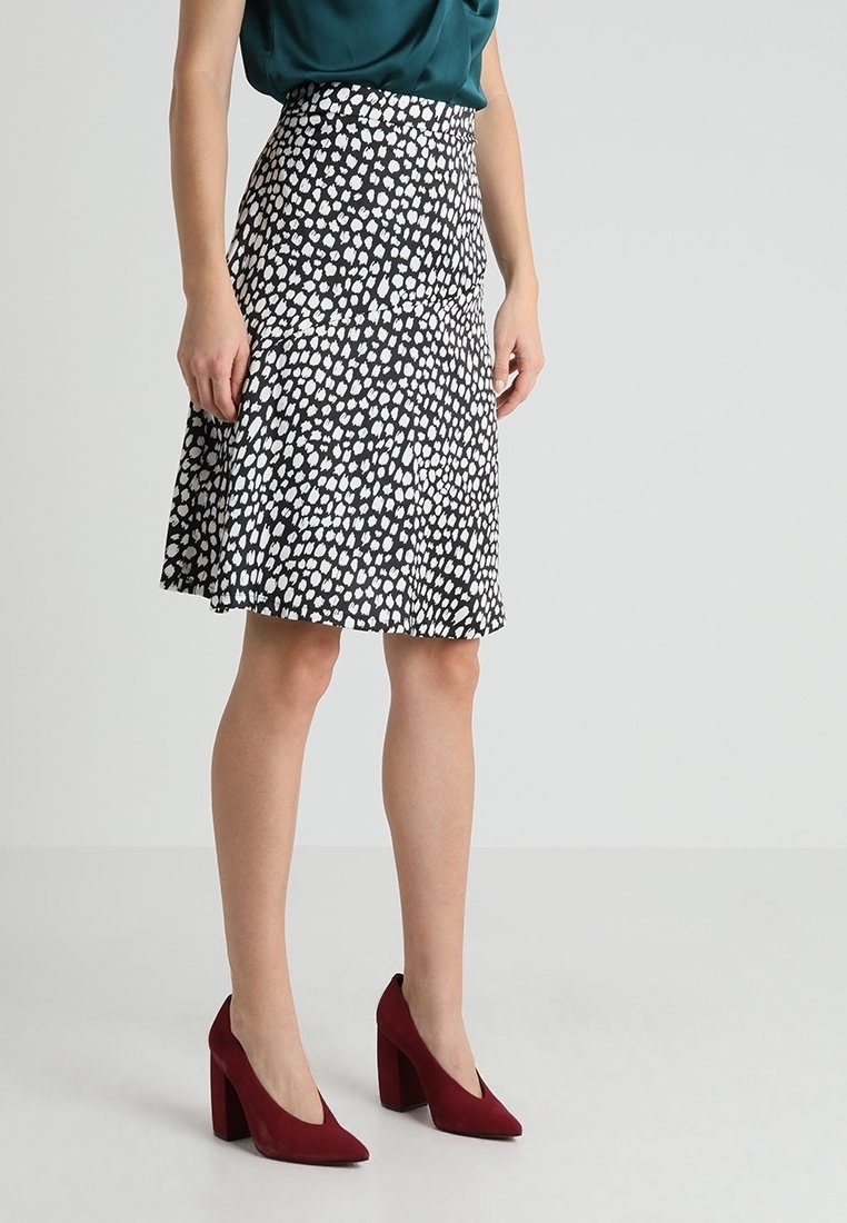Femme portant une jupe mi-longue noire à pois blancs, un haut vert foncé et des chaussures à talons rouges, debout sur un sol clair.