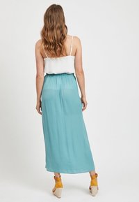 Frau mit welligem Haar, die ein weißes Spaghetti-Träger-Oberteil, einen hellblauen, fließenden Rock und gelbe Keilabsatzsandalen trägt und mit dem Rücken zur Kamera steht.