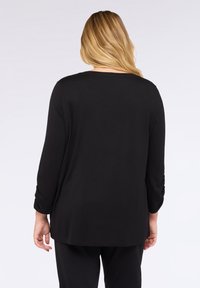 Donna bionda che indossa un semplice top nero a maniche lunghe e pantaloni neri, vista di spalle su uno sfondo bianco puro.