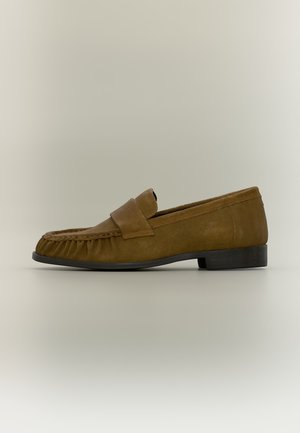 Mocassin en daim marron avec un petit talon noir et une bride sur le dessus, vu de côté sur un fond neutre.