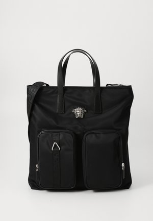 Sac fourre-tout noir en nylon avec des accents en cuir, un logo bien en vue, deux poches zippées à l'avant et une bandoulière amovible.