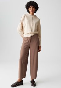 Vrouw met krullend haar die een beige top met lange mouwen, hoge taille wijdvallende bruine broek en donkerbruine instappers draagt, staand tegen een effen achtergrond.