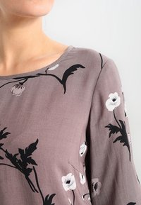 Långärmad blus i dammig mauve-tyg med blommönster i vitt och svart, med rundad halsringning och mjuk textur.