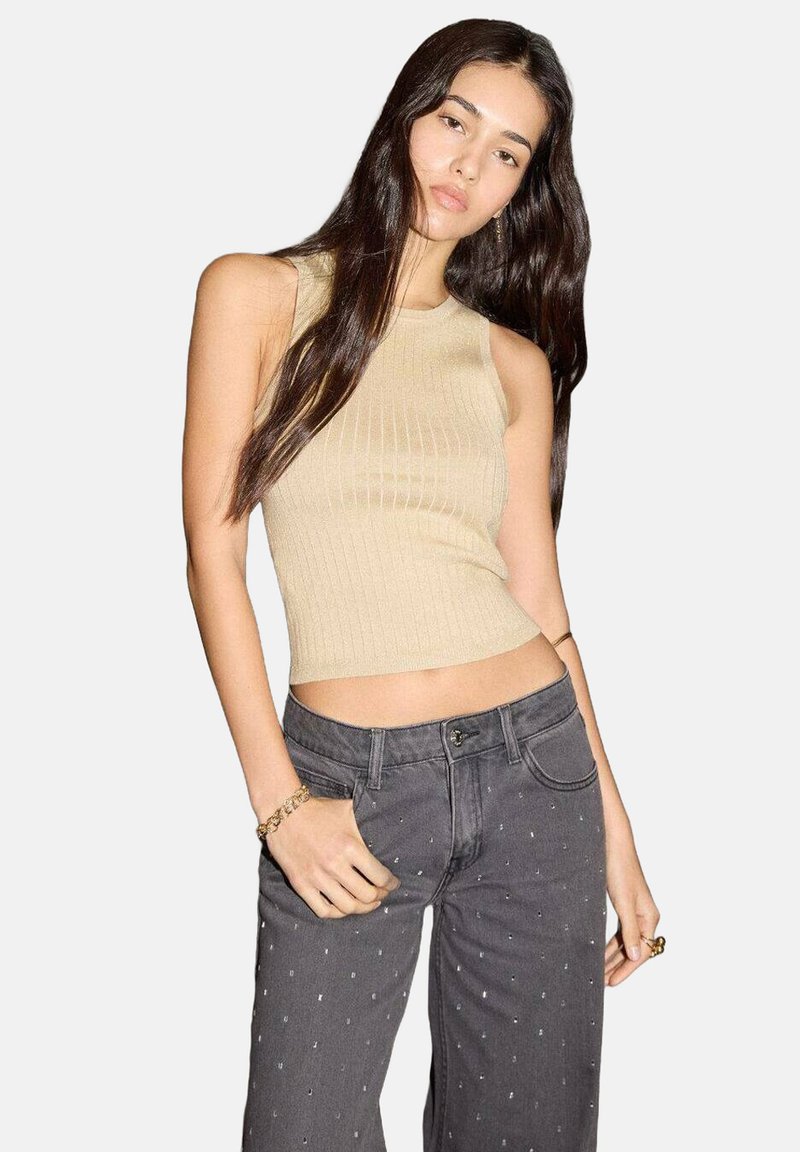 Mladá žena s dlouhými tmavými vlasy má na sobě bezramínkový béžový top s žebrováním a šedé kalhoty s vysok�ým pasem a malými bílými tečkami.