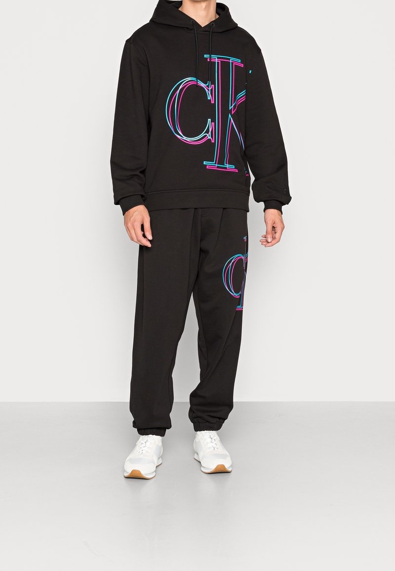 Homme portant un sweat à capuche noir et un jogging avec un grand logo "ck" rose et bleu, associé à des baskets blanches, debout sur un sol gris.