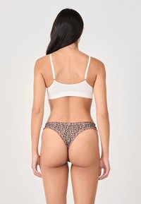 Woman with long dark hair wearing a white bra and leopard print thong underwear with red trim, standing against a plain background.