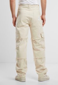 Pantaloni cargo bej din denim cu accente desfăcute, prevăzuți cu buzunare aplicate, o croială lejeră și un detaliu logo pe spate.