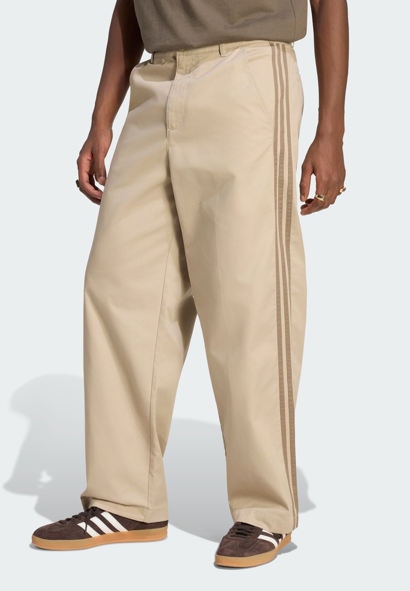 Person wearing beige wide-leg pants with triple side stripes and brown sneakers with white stripes, standing against a plain background.