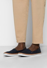 Person wearing beige pants and navy slip-on shoes with white soles and brown trim, standing on a gray floor against a white wall.