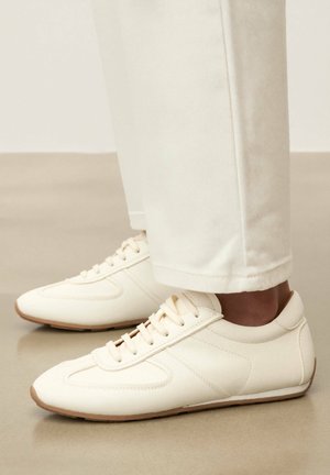 Valged nahast madala säärega tossud beežide talladega, kandes kreemvalgeid sirge säärega pükse siledal põrandal.