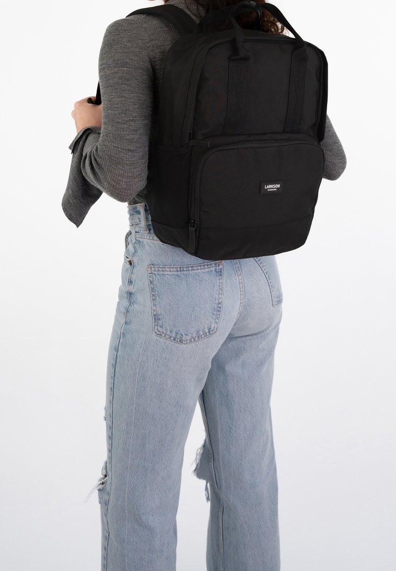 Svart ryggsäck med två handtag upptill och vadderade axelremmar. Huvudfack med dragkedjelås, framficka och märkesetikett.