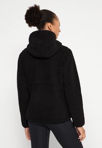 Giacca in pile nera con cappuccio, caratterizzata da una superficie texturizzata, maniche ampie e una vestibilità aderente. Nessun hardware o dettaglio visibile.