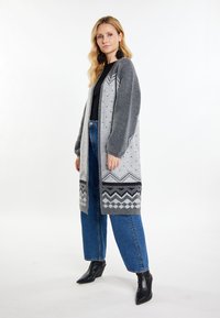 Lungo cardigan grigio a maglia con motivi geometrici, maniche grigio scuro a contrasto e sottili punti decorativi. Indossato sopra un top nero e jeans.