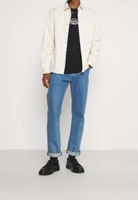 Camisa blanca de botones sobre una camiseta gráfica negra, combinada con jeans azules remangados y zapatillas negras. Diseño simple, ajuste casual.