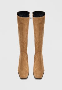 Une paire de bottes hautes en suède beige avec des bouts carrés et des zips sur les côtés, se tenant debout sur un fond blanc.