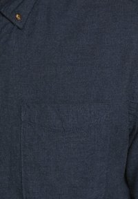 Námořnická modrá košile vyrobená z texturované látky s límcem na knoflíky a jednou náprsní kapsou s viditelnými švy.