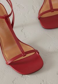 Sandali con tacco in pelle rossa caratterizzati da una punta quadrata, fasce sottili e una superficie liscia. La suola è beige con sottili dettagli di cucitura.