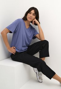 T-shirt bleu à col en V en tissu doux, associé à un pantalon noir court et des chaussures de sport gris clair, posé sur un piédestal blanc.