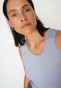 Svetlomodrý rebrovaný top bez rukávov s zaobleným výstrihom a širokými ramienkami. Modelka má rovné, stredne dlhé tmavé vlasy a minimálny make-up.