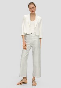 Weißes, kurzes Blazer über einem weißen Tanktop, kombiniert mit hochgeschnittenen, hellgrauen, weiten Jeans und flachen Sandalen mit einem dünnen Riemendesign.