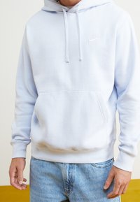 Ljusblå bomullshoodie med justerbara dragsnören, känguruficka och broderad vit Nike-logga på bröstet. Mjuk, borstad textur.