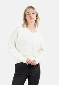 Pulover alb, texturat, cu decolteu în V și mâneci lungi; are un tiv și manșete cu striații, purtat peste pantaloni negri, ajustați.