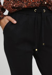 Zwarte taps toelopende broek van zacht stof, met een elastische tailleband met trekkoord en gouden accenten, en zijzakken.