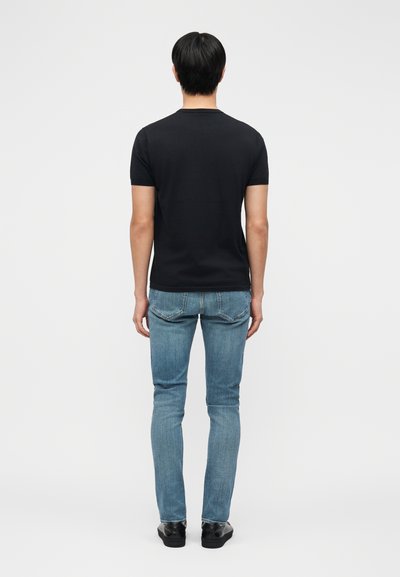 Άνδρας όρθιος με την πλάτη προς τα πίσω, φοράει απλό μαύρο μπλουζάκι με κοντά μανίκια, ανοιχτό μπλε παντελόνι τζιν και μαύρα παπούτσια σε λευκό φόντο.