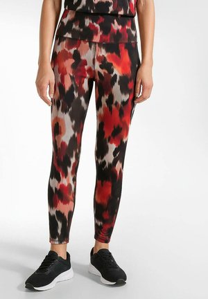 Frau, die hochtaillierte Leggings mit rotem, schwarzem und beigem abstraktem Muster und schwarzen Sportschuhen trägt, steht vor einem weißen Hintergrund.