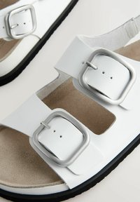 Sandali bianchi con due larghe cinghie, fibbie argentate, un plantare contournato e suola in gomma nera. Morbido effetto scamosciato all'interno.