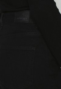 Os jeans de denim pretos apresentam uma textura suave, um corte clássico e uma etiqueta com a inscrição "jeans perfeitos" cosida na cintura.