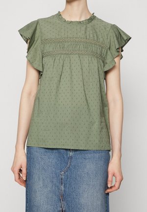 Vrouw die een groene, gestructureerde blouse met korte mouwen en ruches draagt, gecombineerd met een blauwe denim rok.