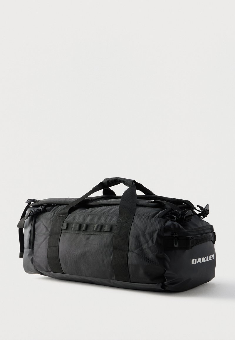 Sort Oakley duffel taske med dobbelthåndtag, sidelommer og lynlåslommer placeret på en hvid baggrund.