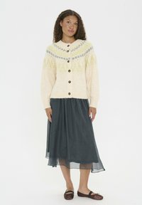 Cardigan de culoare crem cu un model geometric în verde și gri, având nasturi din lemn, manșete dantelate și o textură bulionată, asortat cu o fustă închisă la culoare.