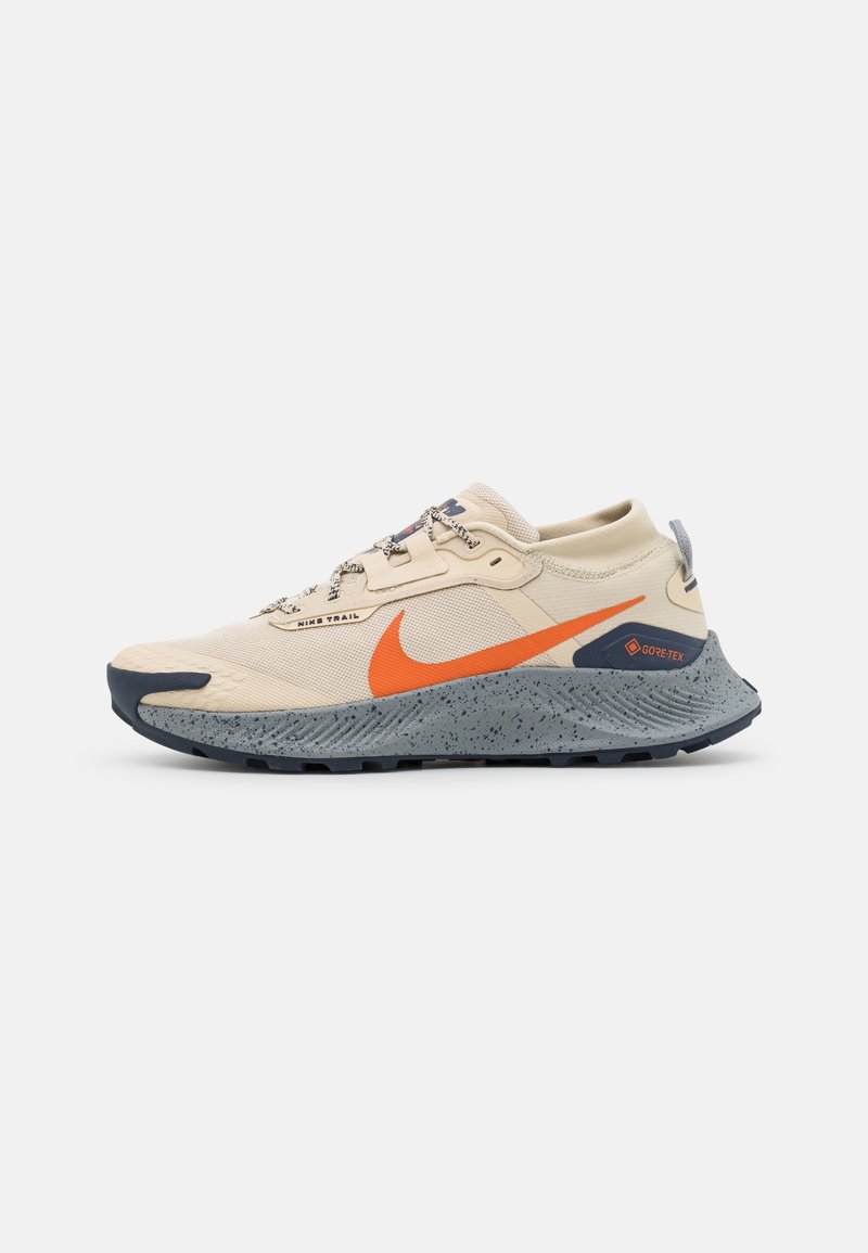 Chaussure de trail Nike beige et grise avec logo Swoosh orange, semelle tachetée et marque Gore-Tex sur le talon.