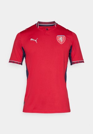 Crvena kratkih rukava dres od prozračnog materijala, s tamnoplave bočne ploče, ovratnikom s gumbom i oznakom grba na prsima.