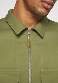 Veste vert olive à fermeture éclair avant, dotée d'un col pointu et de deux poches poitrine. Le tissu est lisse avec des détails de couture visibles.