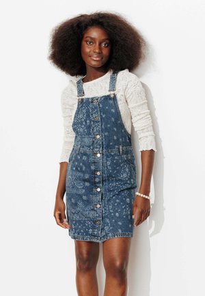 Jeune femme aux cheveux naturels volumineux portant un haut blanc texturé à manches longues et une robe salopette en denim bleue à motif boutonnée.