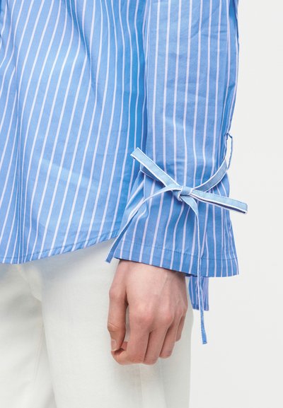 Μανίκι πουκάμισου με κατακόρυφες ρίγες σε μπλε και λευκό, με λεπτομέρεια φιόγκου δεμένη κοντά στον καρπό, φορεμένο με λευκό παντελόνι.