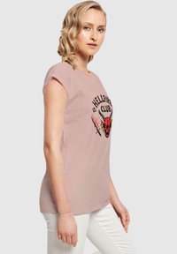 Camiseta rosa clara de manga corta con un diseño gráfico en negro y rojo de un skull con cuernos y el texto "HELLFIRE CLUB."