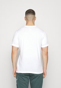 Vit kortärmad t-shirt med rund halsringning, med en enfärgad design och mjuk textur, kombinerad med turkosa byxor. Bakvy.