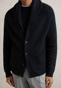 Hombre vistiendo un cárdigan de punto acanalado en azul marino oscuro con cuatro botones y dos bolsillos frontales sobre una camisa negra y pantalones grises.
