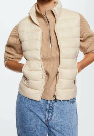 Personne portant une doudoune beige par-dessus un pull côtelé beige avec une fermeture éclair à l'avant, associée à un jean bleu taille haute, les mains posées sur les hanches.