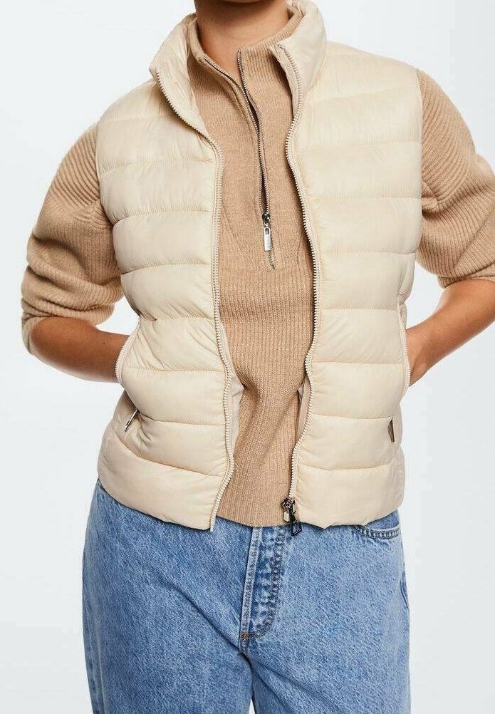 Personne portant une doudoune beige par-dessus un pull côtelé beige avec une fermeture éclair à l'avant, associée à un jean bleu taille haute, les mains posées sur les hanches.