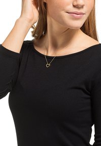Guld hjerteanheng necklace på en tynd kæde, båret over en sort ribbet langærmet bluse. Anhængeren er omridset og enkel i design.