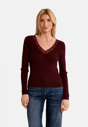 Donna bionda che indossa un maglione a coste bordeaux a maniche lunghe con scollo a V in pizzo e jeans blu, in piedi davanti a uno sfondo chiaro e semplice.