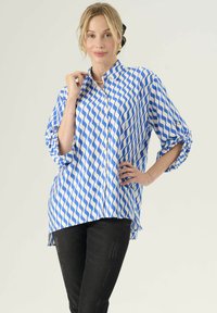 Niebieska i biała koszula z wzorem fal w stylu button-up, z podwiniętymi rękawami i zaokrąglonym dołem, wykonana z lekkiego materiału. W zestawie z czarnymi spodniami.