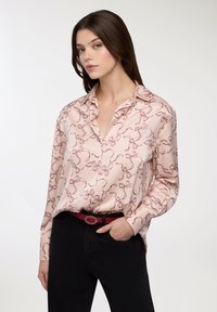 Blusa rosa chiaro con motivo a fiocco, maniche lunghe, colletto e vestibilità morbida. Abbinata a pantaloni neri e una cintura rossa.