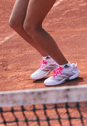 Jambes d'un joueur de tennis portant des chaussures blanches Adidas avec des lacets roses, debout sur un court en terre battue près du filet.
